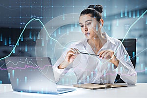 Attractive european businesswoman at desktop using laptop with abstract glowing forex graph on blurry office interior background