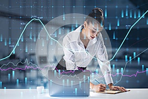 Attractive european businesswoman standing at desktop using laptop with abstract glowing forex graph on blurry office interior