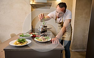 Attractive European male chef decorating spaghetti Bolognese with fresh greens, standing at kitchen island in the home