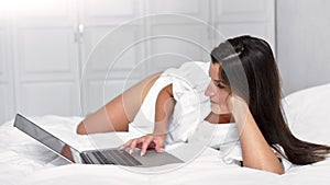 Attractive European woman enjoying weekend lying on bed in bedroom and using laptop touchpad