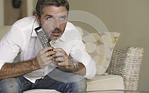 Attractive excited and nervous man watching television in intense concentrated face expression holding TV remote sitting on living