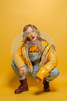 Attractive fashion model woman in winter jacket and sunglasses posing on bright yellow background