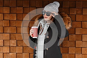 Attractive fashionable young woman hipster in sunglasses in a knitted hat in a winter jacket with a fur hood stands outdoors