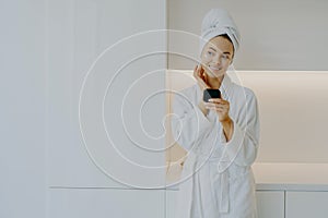 Attractive female applies moisturising cream on face takes care of her complexion and skin holds mirror wears white bathrobe