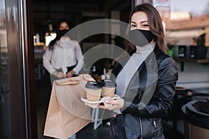 Attractive female customer in face mask standing outdoors by cafe and take away food and coffee. Background of cashier