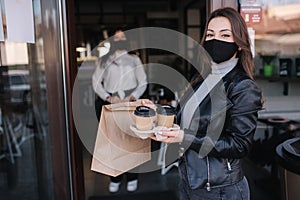 Attractive female customer in face mask standing outdoors by cafe and take away food and coffee. Background of cashier