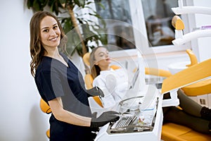 Attractive female dentist doctor standing in her office, with a female patinet in the chair