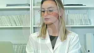Attractive female doctor in glasses using laptop sitting at reception desk