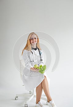 An attractive female doctor in a white coat is sitting smiling and holding an apple and salad in her hands. A cheerful