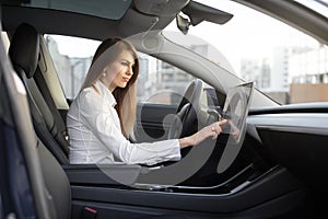 Attractive female driver in formal wear, operating the touchscreen on the dashboard in her electric car inputting data