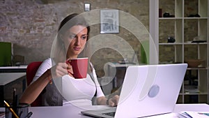 Attractive female office worker typing on the laptop drinking coffee gets statisfied and smiles being on thec workplace