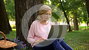 Attractive female pensioner sitting in park with laptop and surfing the net, app