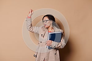 Attractive female professor in beige coat and glasses, having fun, holding a book, singing and dancing on cream backdrop