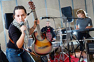 Attractive female soloist playing guitar and singing with her music band in sound studio