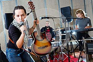 Attractive female soloist playing guitar and singing with her music band in sound studio photo
