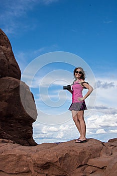Attractive fit young woman photographer stands on top of a red rock formation in Arches National Pa