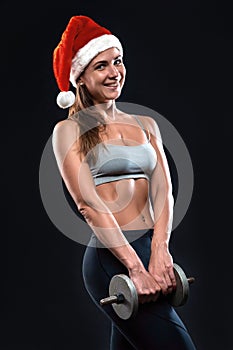 Attractive fitness woman is standing with dumbbells in christmas hat