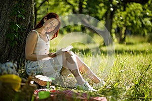 Attractive foxy woman reading book sitting in garden with her back close to tree.