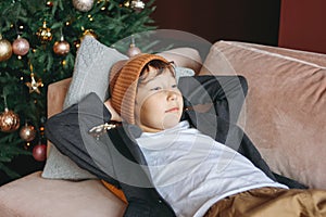 Attractive funny tween boy with dark hair in hat waiting new year on sofa on christmas tree background