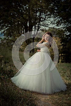 The attractive ginger hair bride with red lipstick in the long wedding dress is posing in the forest.