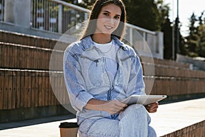 Attractive girl in denim clothes sits on park bench, holds smart tablet in her hands. Portrait of beautiful woman