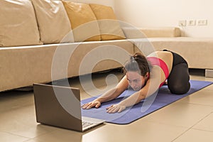 Attractive girl doing yoga on a sports blue rug in sportswear. A laptop with online lessons, she looks at the screen and repeats