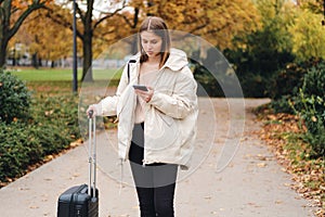 Attractive girl in down jacket with suitcase thoughtfully looking way on cellphone outdoor