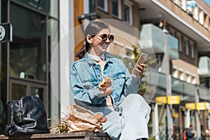 attractive girl eating in the street in the city while using phone to rate the takeaway food company for delicious menu