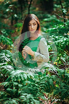 Attractive girl in a green medieval dress sits in the bushes of fern. Village woman herbalist collects herbs for potion in the for