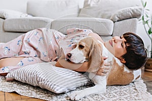 Attractive girl with happy face expression lies on carpet near beagle dog with light-brown ears. Indoor portrait of