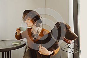 An attractive girl in a hat sits at a coffee table, hugs a dog and drinks coffee. Fashionable lady drinking coffee in an animal
