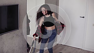 Attractive girl in her 20`s enjoys singing and dancing while vacuum cleaning the apartment.