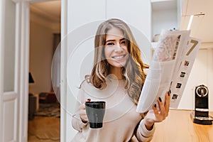 Attractive girl with light-brown hair holding magazine and laughing in her room. Indoor photo of cute brunette woman