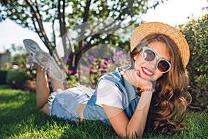 Attractive girl with long curly hair in hat is lying on grass in summer park. She wears jeans jerkin, shorts, sunglasse