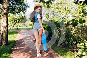 Attractive girl with long curly hair in hat is walking with skateboard in summer park. She wears jeans shorts, jerkin