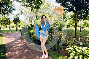 Attractive girl with long curly hair is posing with skateboard in summer park. She wears jeans shorts, jerkin