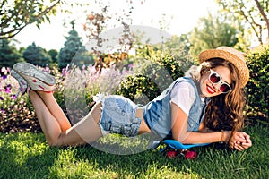 Attractive girl with long curly hair in sunglasses is lying on grass in park. She wears jeans jerkin, shorts, hat. She