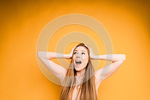 An attractive girl with long hair is surprised and holds her head with both hands on an orange background