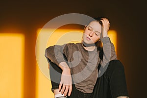 Attractive girl model in casual clothing with eyes closed on orange background, sunshine. Portrait of a woman sitting on an orange