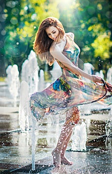 Attractive girl in multicolored short dress playing with water in a summer hottest day. Girl with wet dress enjoying fountains