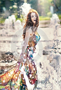 Attractive girl in multicolored short dress playing with water in a summer hottest day. Girl with wet dress enjoying fountains