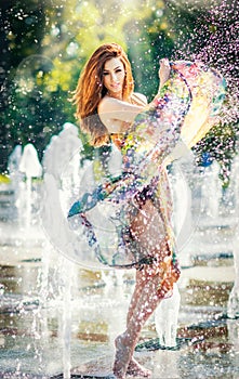 Attractive girl in multicolored short dress playing with water in a summer hottest day. Girl with wet dress enjoying fountains