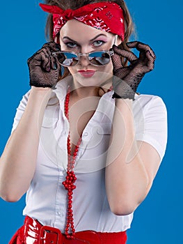 An attractive girl with a red bandana on her head attentively looks with lowered glasses. Girl in pin-up style isolated on blue ba