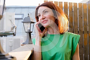 Attractive girl with red hair in a green blouse is talking on a mobile phone on the open terrace of a cafe