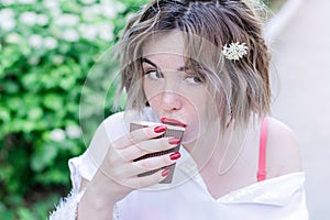 Attractive girl with red lips and manicure whith flower in her hair is sitting in the park and drinking coffee