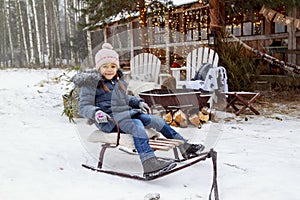 attractive girl rides sleigh in backyard. Snow-covered New Year house with fir trees and hearth with chairs