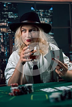 attractive girl in shirt and hat drinking whiskey and looking at poker cards in casino