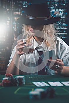 attractive girl in shirt and hat drinking whiskey and looking at poker cards in casino