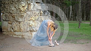 Attractive girl in silver and blue dress puts on high heeles shoes getting ready to pose during photo session near stony