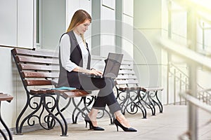 Attractive girl sitting on a bench near a business building with a laptop.